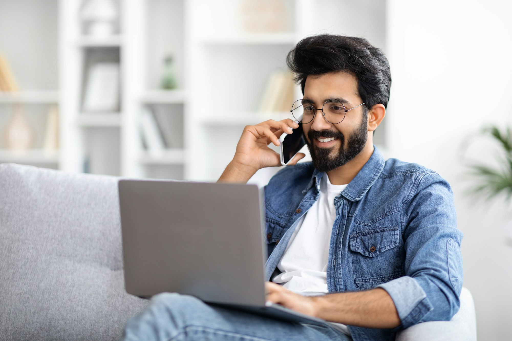 Remote Business. Indian Man Talking On Cellphone And Using Laptop At Home